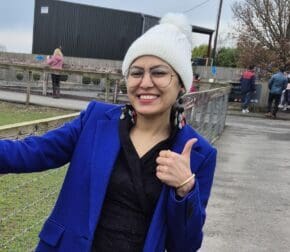 Karu, a young lung cancer patient, smiles and gives a thumbs-up outdoors.