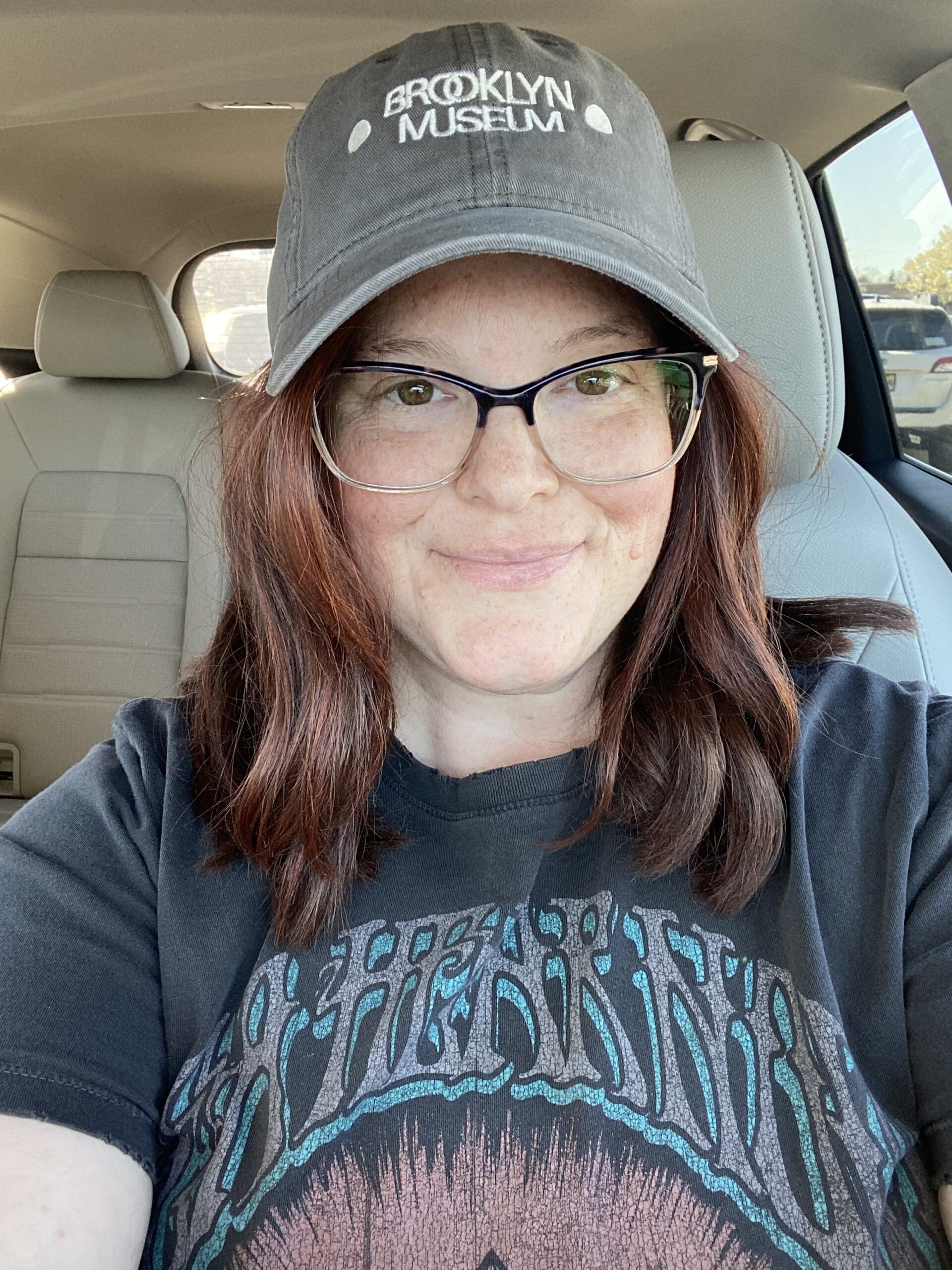 Kelly, a young patient with lung cancer, smiling in a Brooklyn Museum cap while seated in her car.