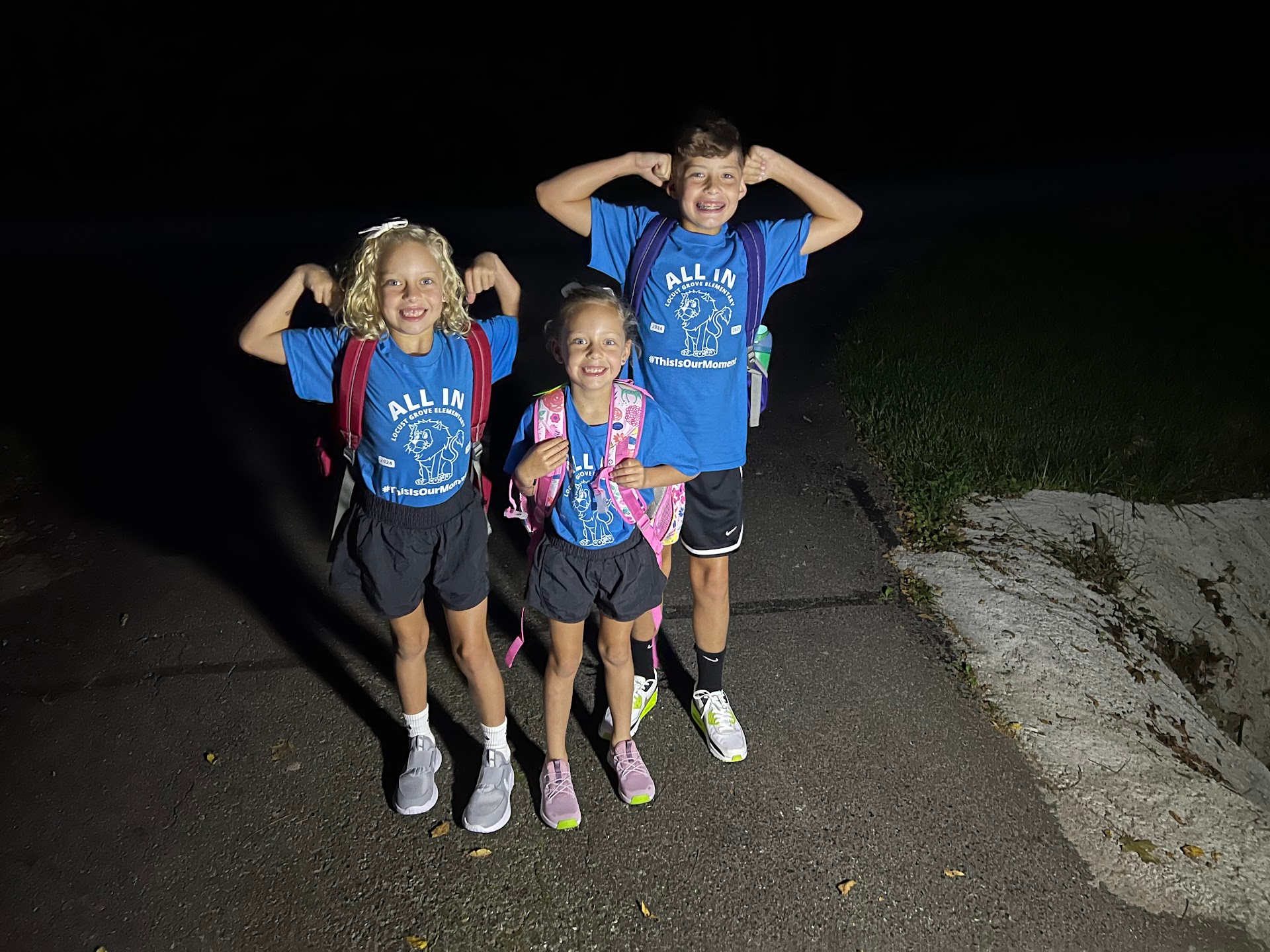 Three of Leah’s children smiling and flexing in matching school shirts with their backpacks