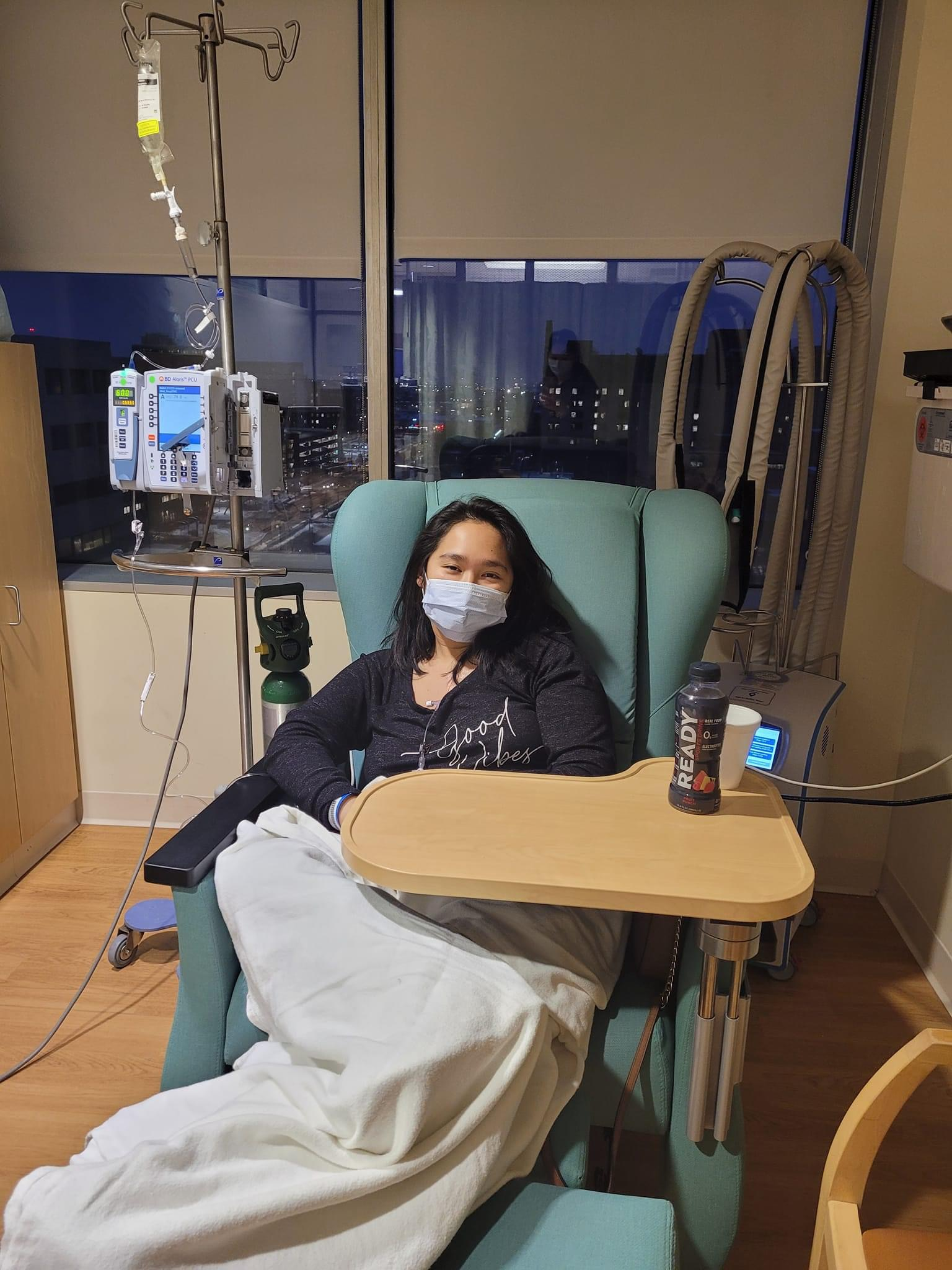Aurora Lucas seated in a clinic chair beside an IV stand.
